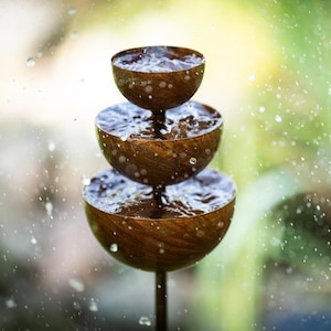 Peut inclure: Une fontaine à eau en bois à trois niveaux, avec de l'eau qui coule de chaque bol. La fontaine a un aspect rustique et naturel, avec des gouttelettes d'eau suspendues dans l'air. L'arrière-plan est flou, avec des teintes vertes et jaunes.