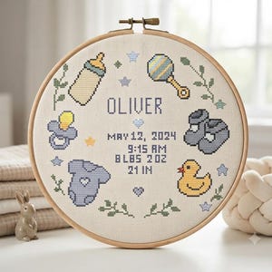 May include: A circular wooden embroidery hoop containing a cross-stitch birth announcement for Oliver. The design features a baby bottle, rattle, pacifier, booties, onesie, and rubber duck in blue, yellow, and green, with birth details included.