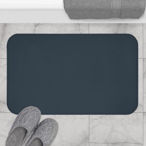 May include: A rectangular dark navy blue bath mat with rounded corners, positioned on a light gray tiled floor next to a pair of gray cable-knit slippers and a folded gray towel, creating a simple and functional bathroom floor accessory.