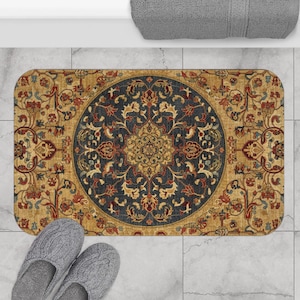 May include: A rectangular bath mat featuring a central dark blue circular medallion with intricate floral and vine patterns in shades of gold, rust red, and cream. The surrounding border is a warm tan color with matching floral motifs.