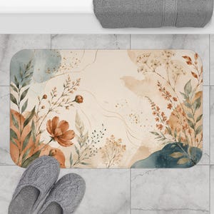 Op de afbeelding: Een rechthoekige badmat met een crèmekleurige achtergrond en botanische illustraties in aquarel van wilde bloemen, bladeren en stengels in tinten oranje, saliegroen en diepblauw, geaccentueerd door subtiele goudkleurige spetters.