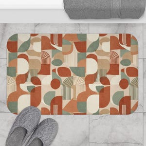 Puede incluir: Una alfombrilla de baño rectangular con un patrón geométrico de formas superpuestas en tonos terracota, verde salvia y crema. El diseño incluye semicírculos, arcos y líneas verticales, creando una composición equilibrada en la superficie.