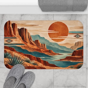 Op de afbeelding: Een rechthoekige badmat met een illustratie van een woestijnlandschap met rode rotsformaties, een grote oranje zon en een blauwe rivier. Het ontwerp bevat horizontale strepen en geometrische ruitpatronen in blauwgroene, oranje en bruine tinten.