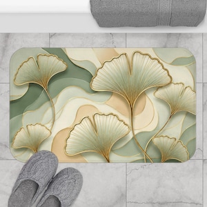 May include: A rectangular bath mat featuring a flowing, abstract pattern of sage green, cream, and beige wavy lines. The design is accented with several large, stylized ginkgo leaves outlined in gold, creating a layered, organic visual effect.