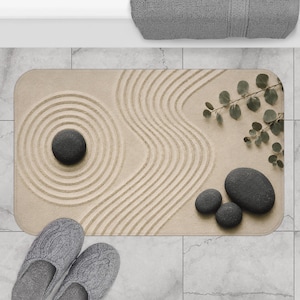 Puede incluir: Una alfombrilla de baño rectangular de color beige con un diseño impreso de arena rastrillada, círculos concéntricos y líneas fluidas. La alfombrilla incluye ilustraciones de cuatro piedras lisas de color gris oscuro y una rama de eucalipto.