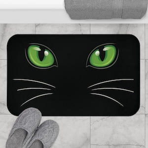 May include: A rectangular bath mat featuring a black background with two large, bright green cat eyes centered in the upper portion. Three thin, white, curved lines extend outward from each side of the mat to represent cat whiskers.