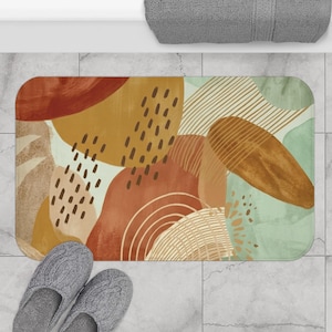 May include: A rectangular bath mat featuring an abstract design with overlapping shapes in shades of terracotta, mustard yellow, and sage green. The pattern includes dark brown dashed lines, fine linear hatching, and concentric semi-circle arcs.