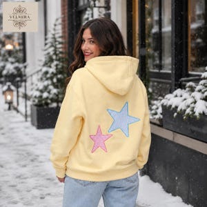 May include: A pale yellow hooded sweatshirt featuring a large light blue and white striped star appliqué and a smaller pink and white checkered star appliqué on the back. The garment has long sleeves, a hood, and a relaxed fit.
