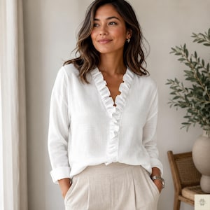 Puede incluir: Una blusa de lino blanco con cuello en V con volantes y mangas largas. La blusa se combina con pantalones de lino beige. La modelo lleva un reloj plateado. La estética general es casual y elegante.