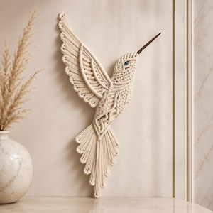 May include: A cream-colored macrame hummingbird wall hanging. The bird has outstretched wings and a long, pointed beak. The eye is a small blue bead. The hummingbird is made of knotted cotton cord.