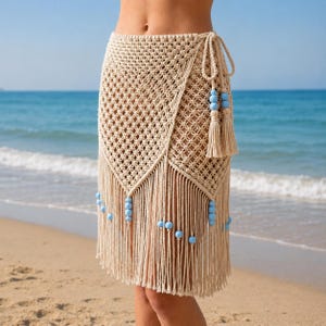 May include: A beige macrame skirt with a wrap design. The skirt features a diamond-patterned weave, fringe detailing, and light blue beads. A tassel hangs from the tie closure. The skirt is worn on a beach.