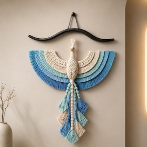 Puede incluir: Un tapiz de macramé con forma de ave con las alas extendidas. La pieza presenta capas de cordón en color crema, azul claro y azul oscuro. Cuelga de un soporte de madera negro curvado. La cola consiste en varias borlas largas de cordón anudado.