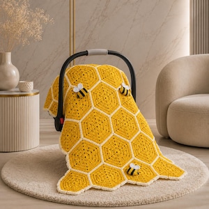 May include: A yellow and white crocheted baby blanket with a honeycomb pattern, adorned with three small bee appliqués. The blanket is draped over a black baby car seat handle, resting on a beige rug.
