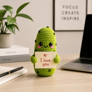 Könnte beinhalten: Eine handgefertigte grüne Häkelkaktusfigur mit schwarzen Augen, rosa Wangen und einem lächelnden Mund. Der Kaktus hält eine kleine Karte mit einem roten Herzen und den Worten "I love you". Im Hintergrund sind ein Laptop, eine Pflanze und ein gerahmter Druck.