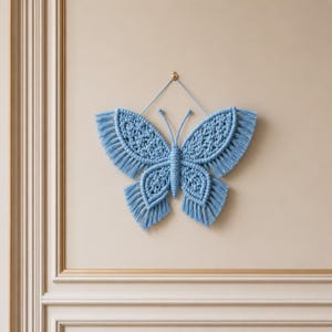May include: A light blue macrame butterfly wall hanging featuring intricate knotted patterns and long, fringed edges on the wings. The butterfly has two thin antennae extending upwards and is suspended from a single small gold-coloured metal hook.