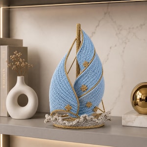 Pode incluir: Um veleiro de crochê azul claro feito à mão com detalhes dourados e enfeites florais. O barco apresenta um design texturizado e detalhado e assenta sobre uma base cinza ondulada. Artigo náutico decorativo.