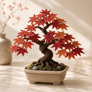 Può includere: Un bonsai all'uncinetto fatto a mano con foglie d'acero rosse e arancioni. Il tronco marrone è attorcigliato, posto in un vaso beige all'uncinetto con muschio verde e piccole pietre. Le foglie sono in varie tonalità di rosso e arancione.