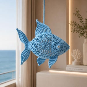 May include: A light blue macrame fish hanging ornament crafted with intricate knotted cord patterns. The design features a prominent dorsal fin, a detailed tail, and a circular eye element, all suspended by a matching blue cord for display.