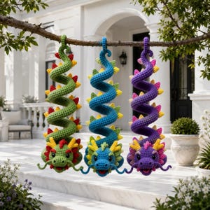 May include: Three crocheted dragon ornaments hanging from a tree branch. The dragons are green, blue, and purple, each featuring a long, spiraling body, small spikes along the back, and detailed faces with whiskers and yellow accents.