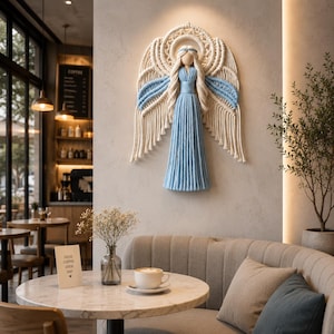 May include: A macrame angel wall hanging with cream and light blue details. The angel has long flowing hair and a long dress. The wings are large and detailed. The angel is hanging on a neutral colored wall in a cafe.