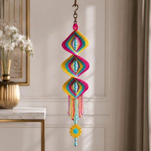 May include: A hanging crochet mobile featuring three diamond-shaped sections in vibrant pink, yellow, and blue yarn. Each section contains a central light blue bead. The piece ends with a yellow crocheted flower and a long, colorful fringe tassel.