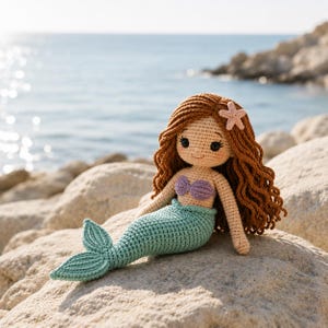 Peut inclure: Poupée sirène au crochet avec de longs cheveux roux et une étoile de mer dans les cheveux. La poupée porte un haut en coquillage violet et une queue turquoise. Elle est assise sur un rocher près de la mer.