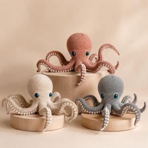 Op de afbeelding: Drie gehaakte octopus speelgoed in beige, koraal en grijs, elk met blauwe ogen en gedetailleerde tentakels. De octopussen worden tentoongesteld op kleine houten sokkels tegen een neutrale achtergrond. Deze handgemaakte pluche speelgoed zijn geschikt voor kinderen.