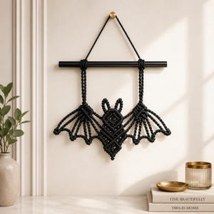 Könnte beinhalten: Schwarze Makramee-Fledermaus-Wanddekoration. Die Fledermaus hat ausgebreitete Flügel und ist an einer schwarzen Stange aufgehängt. Die Fledermaus besteht aus geknotetem schwarzem Seil, und die Flügel haben ein detailliertes, offenes Design.