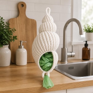 Può includere: Un porta sacchetti a forma di conchiglia, lavorato all'uncinetto, color crema. Il porta sacchetti è riempito di sacchetti di plastica verdi e ha un anello nella parte superiore per appenderlo. Il design ricorda una conchiglia di nautilus. Il porta sacchetti è su un piano di lavoro in legno.