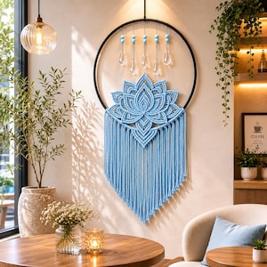 May include: A light blue macrame wall hanging featuring a lotus flower design, suspended within a black metal ring. The piece includes hanging strands and decorative crystal-like accents, creating a bohemian aesthetic. The wall decor is displayed in a well-lit room.