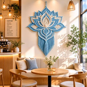 May include: A large, blue and cream-colored macrame lotus flower wall hanging. The intricate design features layered petals and a fringed bottom. The wall hanging is displayed in a cafe setting with a coffee menu visible.