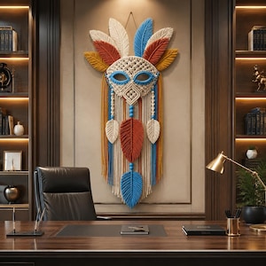 Op de afbeelding: Een macramé wandhanger met een centraal maskerontwerp met blauwe oogaccenten, omringd door crèmekleurige, roestkleurige, blauwe en gele vezels in de vorm van veren. Het stuk bevat lange hangende koorden, decoratieve blauwe kralen en een symmetrische opbouw.