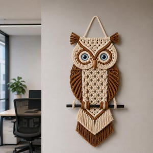 May include: A macrame owl wall hanging in shades of brown and cream. The owl features large blue eyes, a wooden beak, and a decorative fringe at the bottom. The owl is hanging on a black rod.