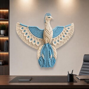 May include: A wall hanging crafted from macrame cord in the shape of a bird with wings spread wide. The design features a combination of cream and light blue knotted patterns, with fringed edges forming the feathers of the wings and tail.