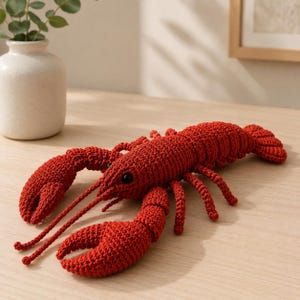 May include: A handmade, red crochet lobster with black eyes and long antennae. The lobster has large claws and multiple legs, and is displayed on a light-colored wooden surface. The lobster is a decorative item.