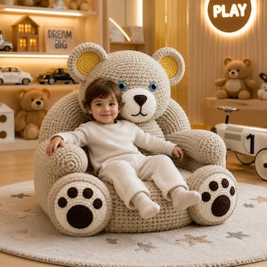 May include: A beige crochet bear-shaped armchair featuring a large head with yellow inner ears, blue eyes, and a dark brown nose. The armrests are designed as bear paws with dark brown circular pads and toes, providing a soft seat for a child.