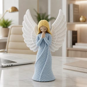 Peut inclure: Une figurine d'ange faite à la main au crochet, avec des ailes blanches, une robe bleu clair et des cheveux blonds. L'ange est en position de prière. La figurine est exposée sur une surface en marbre blanc, avec un ordinateur portable et un cadre de bureau en arrière-plan.