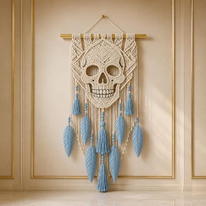 May include: A macrame wall hanging featuring an intricate off-white cord skull design suspended from a gold-toned horizontal rod. The piece is adorned with light blue tassels and several light blue leaf-shaped fibre accents hanging vertically.