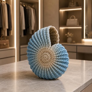 May include: A nautilus shell-shaped decorative object crafted from crochet yarn. The piece features a gradient color pattern transitioning from a light beige center to a vibrant sky blue along the outer ribbed edges, creating a spiral design.