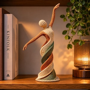 May include: A handcrafted crochet sculpture of a dancer in a graceful pose. The figure features a spiral design with shades of beige, orange, and green. The sculpture is displayed on a wooden shelf, with a book and a lamp in the background.