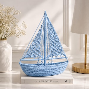 May include: A light blue macrame sailboat model. The decorative nautical piece features a detailed woven design, including the hull, mast, and sails. The sailboat is displayed on a white book titled "The Kinfolk Home".