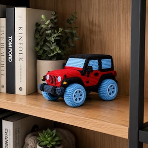 Puede incluir: Un Jeep de juguete tejido a ganchillo en rojo, negro y azul. El Jeep presenta ruedas azules, un techo negro y una carrocería roja. El juguete se exhibe en una estantería de madera con libros y una pequeña planta en el fondo.