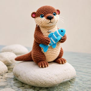 May include: A crocheted otter figurine in brown and beige, holding a blue and white striped fish. The otter has blue eyes, pink ears, and white whiskers. It is sitting on a smooth, gray stone, with water in the background.