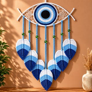 Op de afbeelding: Een macramé wandhanger met een centraal boze oog motief in wit, lichtblauw en donkerblauw garen. Zeven gevlochten koorden hangen aan de basis, elk versierd met een groene kraal en een bladvormig macramé kwastje in een wit naar blauw verloop.