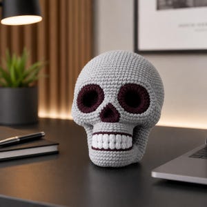 May include: A crocheted skull sculpture in gray with burgundy eye sockets and nose, and white teeth. The skull is a decorative item, possibly for Halloween or gothic decor. The skull is sitting on a dark surface.
