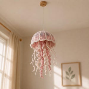 Puede incluir: Lámpara colgante de ganchillo hecha a mano con forma de medusa. La lámpara presenta una combinación de colores rosa y blanco, con una parte superior redonda y abovedada y largos tentáculos fluidos. Se suspende de un soporte de techo de madera.