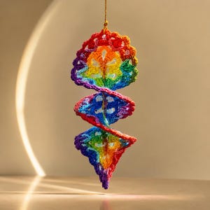 May include: A hanging spiral ornament crafted from multicoloured crochet yarn. The design features a repeating pattern of red, orange, yellow, green, blue, and purple sections arranged in a vertical, twisting structure suspended by a thin gold string.