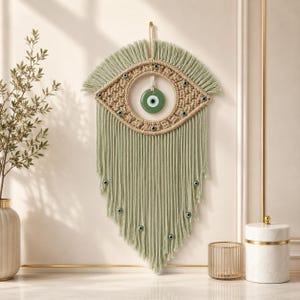 May include: A sage green macrame wall hanging in an eye shape, featuring a central green eye charm and smaller eye charms. The macrame has a woven center and fringed edges, creating a decorative wall art piece.
