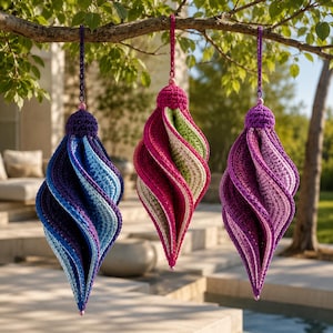 Op de afbeelding: Drie spiraalvormige gehaakte ornamenten hangend aan een boomtak. Het linker ornament heeft tinten blauw en paars, het middelste toont roze, groen en crème, en het rechter ornament laat verschillende tinten paars en lavendel zien.