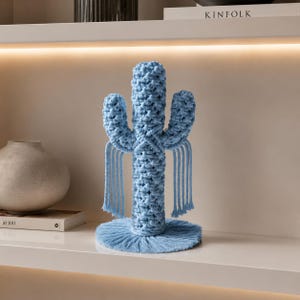 May include: A light blue macrame cactus sculpture featuring a central column with two side arms, a circular base, and long fringe details hanging from each arm. The piece is constructed from knotted cord with a textured, woven appearance.